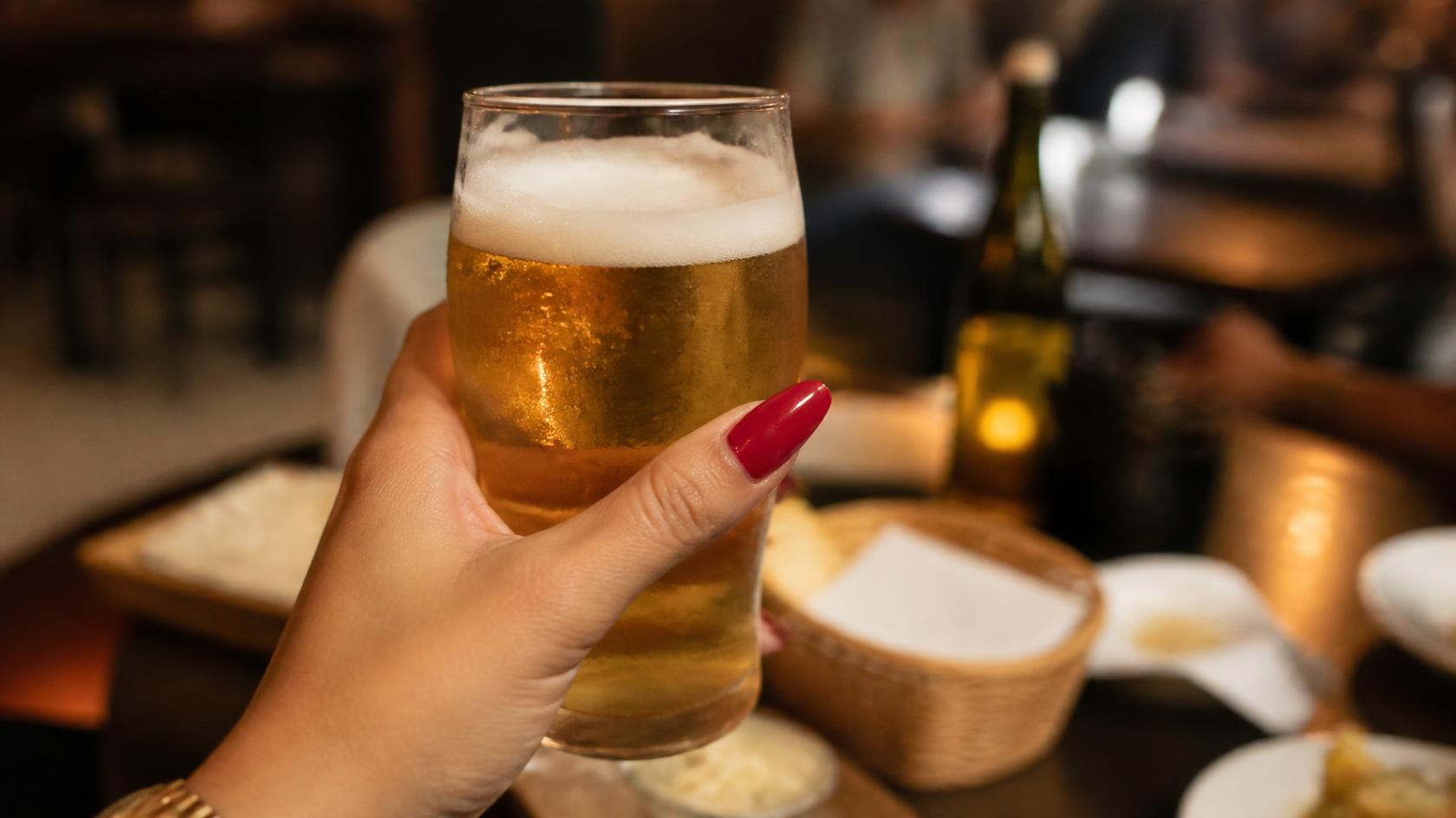 Imagem: Uma mao feminina com esmalte vermelho e unhas longas segurando um copo de cerveja com fundo desfocado de uma mesa de bar fotografia realista foco na mao e no copo atmosfera de bar aconchegante Consumo excessivo de álcool entre mulheres brasileiras dobra nas últimas duas décadas